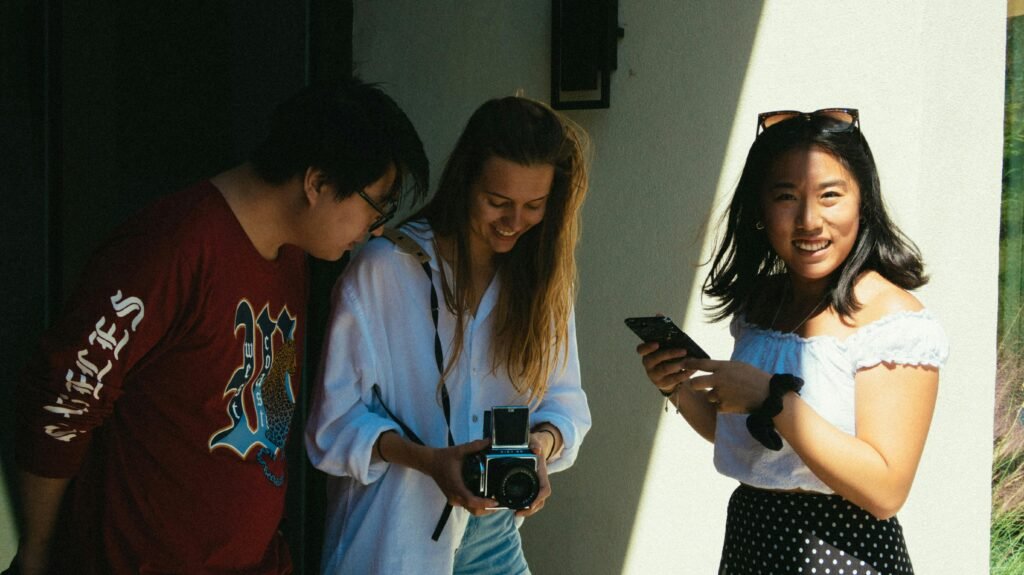 Three young friends smiling, sharing moments with a camera and phone outdoors, enjoying a sunny day.